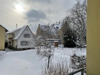 Eine verschneite Landschaft mit Häusern und Bäumen. Der Himmel ist bewölkt, und die Umgebung wirkt ruhig und winterlich.