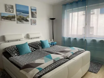 Ein modernes Schlafzimmer mit einem großen Bett und bunten Kissen. Die Wände sind mit Bildern dekoriert, und ein Fenster mit Vorhängen lässt viel Licht herein.