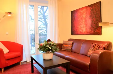 Ein gemütliches Wohnzimmer mit einem braunen Sofa und einem roten Sessel. Eine Pflanze steht auf einem schwarzen Tisch, und große Fenster lassen viel Licht herein.