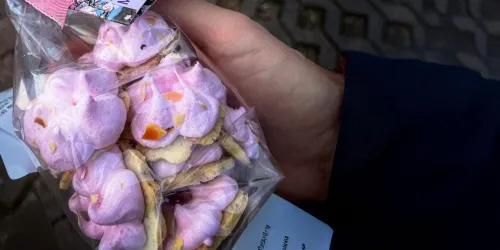 Eine Hand hält eine Tüte mit pinken Süßigkeiten, die wie kleine Wolken aussehen. Die Leckereien haben eine cremige Textur und sind mit bunten Stückchen verziert.