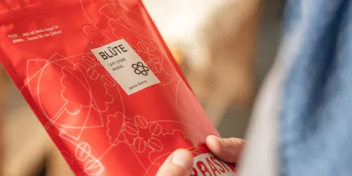 Eine Person hält eine rote Verpackung mit dem Aufdruck "BLÜTE" in der Hand. Die Verpackung zeigt eine moderne, grafische Gestaltung.