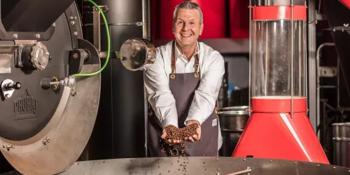 Ein Mann in einer Kaffeerösterei hält frisch geröstete Kaffeebohnen in den Händen. Im Hintergrund sind Röstanlagen und Ausstattungen zu sehen.