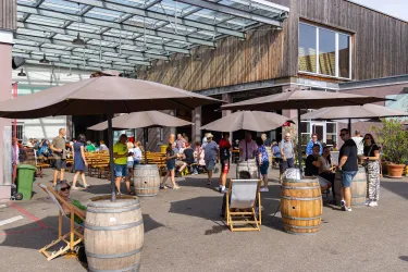 Ein belebter Außenbereich mit Tischen und Sonnenschirmen. Viele Gäste genießen die Zeit in einem entspannten Ambiente.