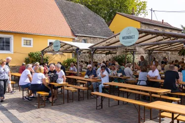 Eine lebhafte Außensitzfläche mit vielen Gästen, die an langen Tischen sitzen. Im Hintergrund sind bunte Gebäude und grüne Pflanzen zu sehen.
