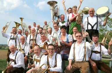 Eine Musikkapelle mit vielen Mitgliedern in traditioneller Tracht. Sie posieren fröhlich im Freien mit verschiedenen Instrumenten.