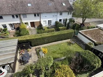 Ein gepflegter Garten mit einem grünen Rasen und farbigen Pflanzen. Im Hintergrund sind Wohnhäuser und eine gepflegte Hecke zu sehen.