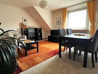 Ein modernes Wohnzimmer mit einem großen Tisch und schwarzen Stühlen. Es gibt eine gemütliche Sitzecke und viel Tageslicht durch ein großes Fenster.