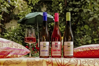 Drei Flaschen Wein stehen auf einem Tisch in einem schönen Garten. Ein Weinglas und farbige Kissen verleihen der Szenerie eine entspannte Atmosphäre.