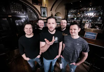 Eine fünfköpfige Band steht lächelnd in einer Bar vor einer Theke mit Getränken. Die Atmosphäre ist entspannt und freundlich.