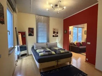 Ein gemütliches Schlafzimmer mit einem Doppelbett und bunten Kissen. Im Hintergrund befindet sich eine Sitzecke mit einem Sofa und Bildern an der Wand.