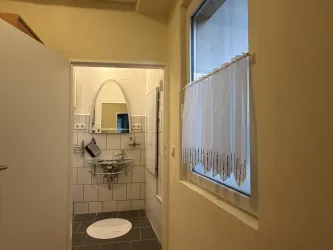 Ein kleines Badezimmer mit hellen Fliesen und einer großen Spiegelwand. Ein Fenster mit einer Vorhang aus weißem Stoff sorgt für natürliches Licht.