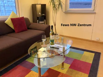 Ein gemütlicher Wohnraum mit einem braunen Sofa und einem bunten Teppich. Auf dem Glastisch stehen Weingläser und eine Flasche Wein.