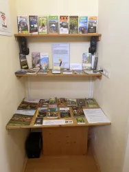 Ein kleiner Informationsstand mit verschiedenen Broschüren auf zwei Regalen. Der Tisch ist mit weiteren schriftlichen Materialien und Informationsblättern bedeckt.