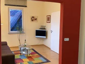 Ein gemütliches Wohnzimmer mit einem braunen Sofa und einem bunten Teppich. An der Wand hängt ein Fernseher und es gibt eine Pflanzen sowie ein paar Bilder.