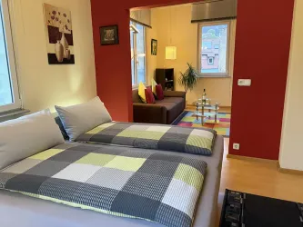 Ein gemütliches Zimmer mit einem Doppelbett und bunten Kissen. Im Hintergrund befindet sich ein Wohnzimmerbereich mit einer Couch und einem bunten Teppich.