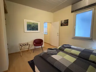 Ein einfaches Schlafzimmer mit zwei Betten und einer roten Stuhl. An der Wand hängen Bilder, und ein Fenster lässt Tageslicht herein.