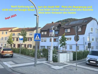 Ein Straßenbild mit mehreren Häusern in einer Stadt. Es sind neue Baustellen und Verkehrsschilder sichtbar, während der Himmel klar und blau ist.