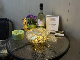 Ein runder Tisch mit einem glitzernden Deko-Element, einer Flasche Wein und einem kleinen Pflanzenkübel. daneben befindet sich ein menürahmen und ein grüner Aschenbecher.