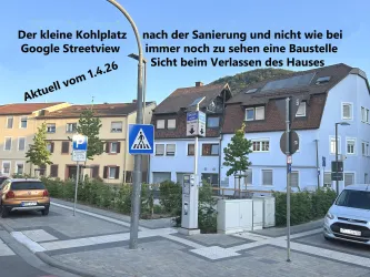Ein kleiner Platz mit moderner Bebauung und begrünten Flächen. Im Vordergrund sind Parkplätze und Verkehrsschilder zu sehen.