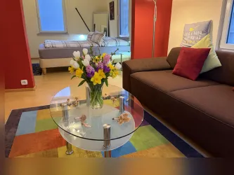 Ein gemütliches Wohnzimmer mit einem braunen Sofa, einem Glastisch und frischen Blumen. Im Hintergrund ist ein Schlafzimmer mit einem Bett und einem Fenster sichtbar.