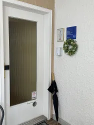 Eine weiße Tür mit einem transparenten Fenster und einem kleinen Kranz ist zu sehen. Neben der Tür befindet sich ein rechtwinkliger Bereich mit einem Schirm und einem Hinweisschild.