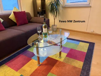 Ein gemütliches Wohnzimmer mit einem dunklen Sofa und mehreren bunten Kissen. Auf einem runden Glastisch stehen eine Flasche Wein und Gläser neben einem bunten Teppich.