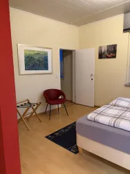Ein gemütliches Schlafzimmer mit einem Bett, einem roten Stuhl und einem kleinen Tisch. An den Wänden hängen Bilder, und eine Tür führt in einen weiteren Raum.