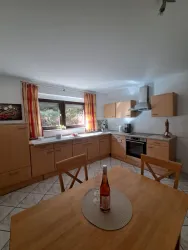 Eine helle Küche mit Holzmöbeln und einer gemütlichen Essgruppe. Durch die Fenster scheint Tageslicht und es gibt einige Küchenutensilien auf der Arbeitsfläche.