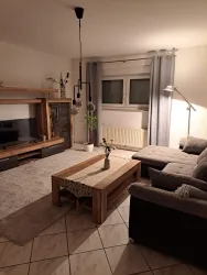 Ein gemütliches Wohnzimmer mit einem grauen Sofa und einem Holztisch. Im Hintergrund befindet sich ein Fernsehschrank und große Fenster mit Vorhängen.