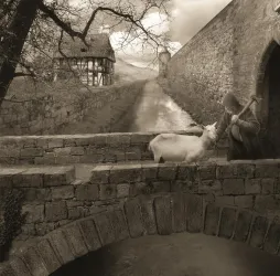 Eine sepiafarbene Landschaft mit einer alten Steinbrücke und einem Hirten, der ein Schaf streichelt. Im Hintergrund sind ein historisches Haus und eine Mauer zu sehen.