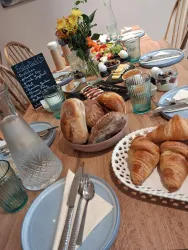 Ein liebevoll gedeckter Frühstückstisch mit frischen Brötchen, Croissants und einer bunten Obstschale. Im Hintergrund steht eine Tafel mit Frühstücksoptionen.