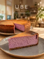 Ein Stück lila Ube-Käsekuchen auf einem grauen Teller. Im Hintergrund sieht man einen weiteren Käsekuchen und eine gemütliche Café-Atmosphäre.