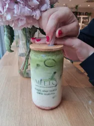 Ein Glas mit Matcha-Getränk und einer Holzkappe steht auf einem Holztisch. Im Hintergrund sind Blumen in einer Vase sichtbar.