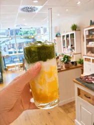 Ein erfrischendes Getränk in einem hohen Glas, mit Schichten aus gelbem Fruchtsaft und grünem Tee. Im Hintergrund ist ein gemütliches Cafe mit Pflanzen und Dekorationen zu sehen.