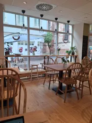 Ein gemütliches Café mit Holztischen und Stühlen. Große Fenster bieten einen Blick auf den Außenbereich mit Pflanzen und Dekorationen.