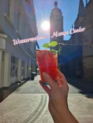 Ein erfrischender Wassermelonen-Mango-Cooler wird in einer Hand gehalten. Im Hintergrund ist eine sonnige Straße mit historischen Gebäuden zu sehen.