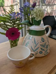 Eine schöne Teekanne mit einem floralem Muster steht neben einer Tasse. Im Hintergrund sind frische Blumen in einer Vase zu sehen.