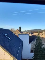 Ein Blick auf ein Wohngebiet mit verschiedenen Dachformen und einer klaren blauen Himmel. Im Hintergrund sind sanfte Hügel und Bäume zu sehen.