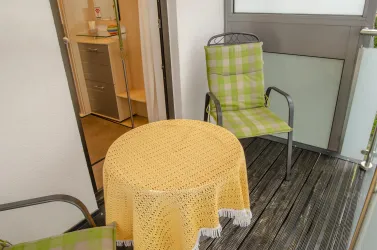 Ein gemütlicher Balkon mit zwei gepolsterten Stühlen und einem runden Tisch. Der Tisch ist mit einer gelben Deckchen bedeckt und die Umgebung wirkt einladend.