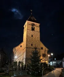 Eine alte Kirche mit einem hohen Turm steht im Dunkeln. Einige Lichter und ein Weihnachtsbaum sind im Vordergrund sichtbar.