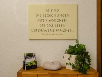 Ein Wandbild mit einem Zitat von Guy de Maupassant über die Wichtigkeit menschlicher Begegnungen. Neben dem Bild steht ein blühender Topf und eine kleine Holzkommode.