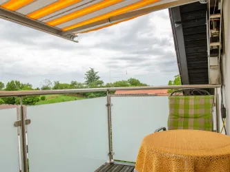 Ein gemütlicher Balkon mit einem runden Tisch und einem Stuhl. Im Hintergrund ist eine grüne Landschaft unter einem bewölkten Himmel zu sehen.