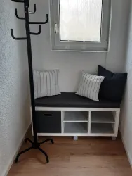 Ein kleiner, heller Flur mit einer Sitzbank und Kissen. An der Wand steht ein schwarzer Kleiderständer.