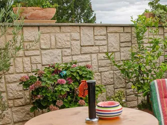 Ein gemütlicher Gartenbereich mit einem Holztisch und bunten Tellern. Im Hintergrund sind Pflanzen und eine Steinmauer zu sehen.