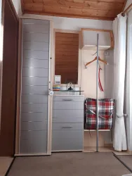 Ein kleines Zimmer mit einem grauen Schrank, einer Kommode und einem Tisch. An der Wand hängt ein Kleiderständer und ein kariertes Sitzkissen ist zu sehen.