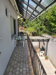 Ein sonniger Balkon mit Fliesen und einem Tisch sowie zwei Stühlen. Im Hintergrund sieht man grünes Laub und einen Bereich mit Pflanzen.