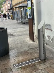 Eine moderne Trinkwasserstation auf einem gepflasterten Gehweg. Im Hintergrund sind Tische und Stühle in einem Café sichtbar.