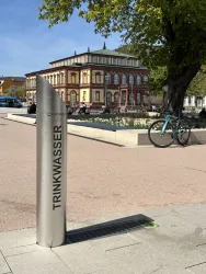 Ein Trinkwasserbrunnen aus Edelstahl steht in einer Stadtlandschaft. Im Hintergrund ist ein historisches Gebäude und einige Bäume zu sehen.