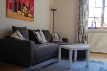 Ein komfortables Wohnzimmer mit einem grauen Sofa und dekorativen Kissen. Ein weißer Tisch steht vor dem Sofa, und ein Fenster lässt viel Tageslicht herein.