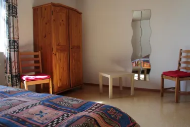 Ein einfaches Zimmer mit einem Holzschrank, zwei Stühlen und einem kleinen Tisch. Im Hintergrund ist ein verzerrter Spiegel zu sehen.